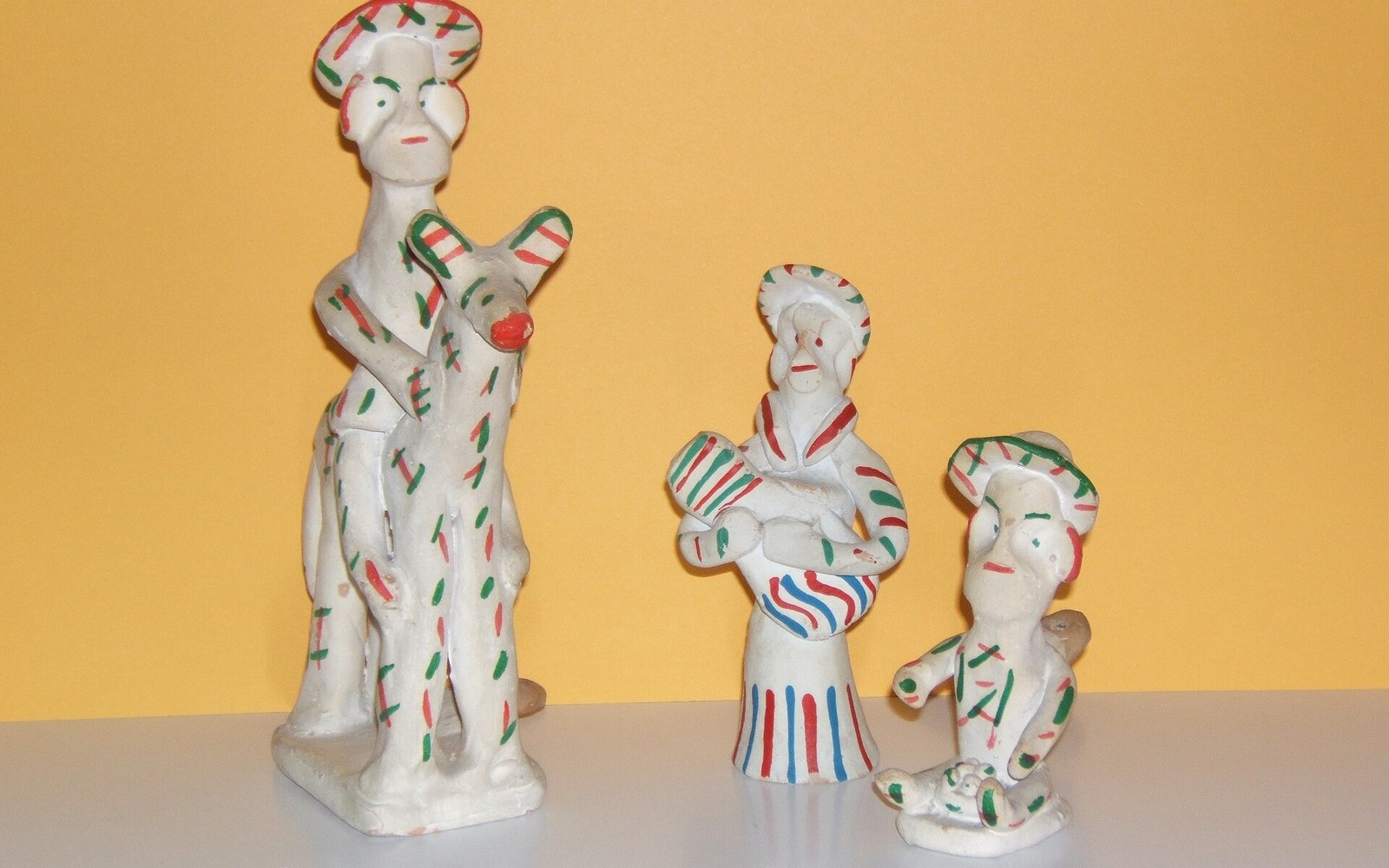 Three white clay siurell figurines painted with red and green stripes stand against an orange backdrop