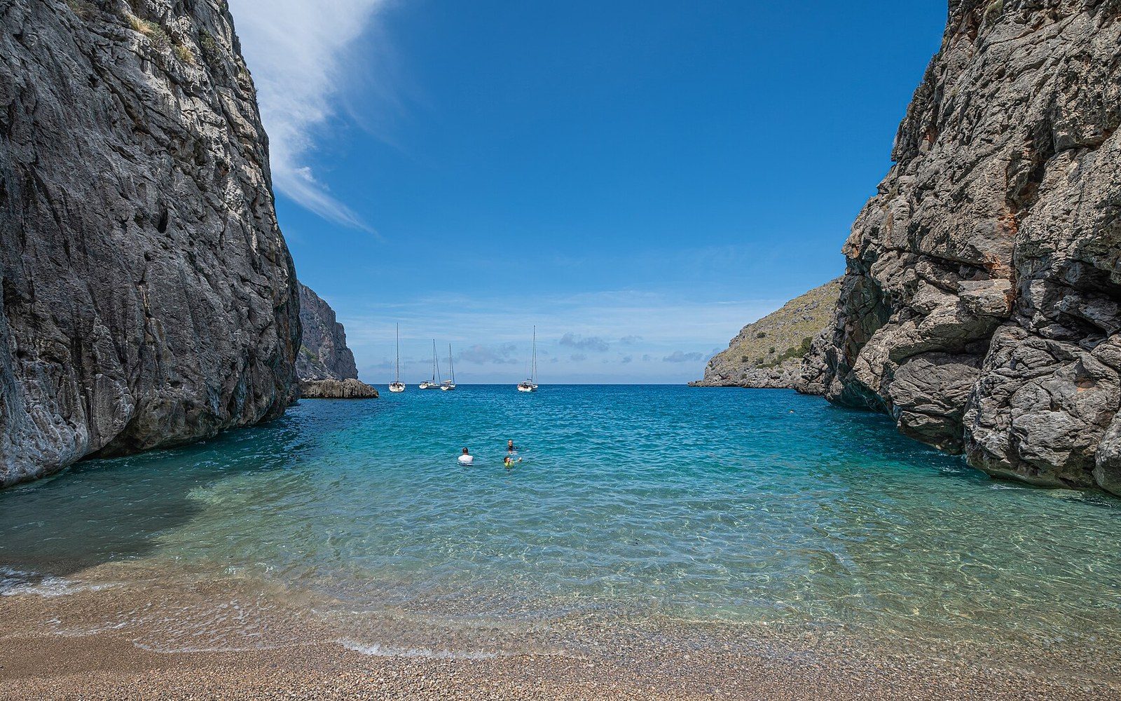 Swimmers wade in a turquoise cove between towering limestone cliffs, sailboats anchored offshore